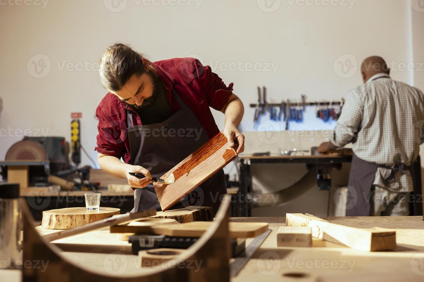 Carpenter brushing varnish on wood to build up protective layer with african american coworker in background. Woodworker lacquering plank after sanding surface next to BIPOC apprentice working photo