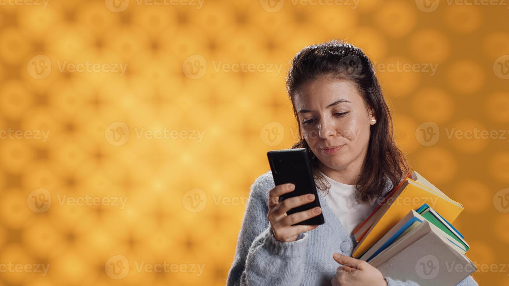 Smiling woman reading messages on smartphone while holding stack of books, isolated over studio background. Cheerful person with pile of academic textbooks in arms texting friends on phone, camera A photo