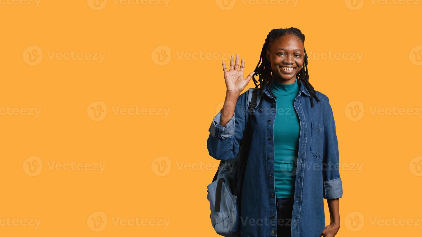 Portrait of chipper girl holding backpack, waving hand, gesturing, isolated over yellow studio background. Jolly african american student with school rucksack doing greeting hand gesture, camera B photo