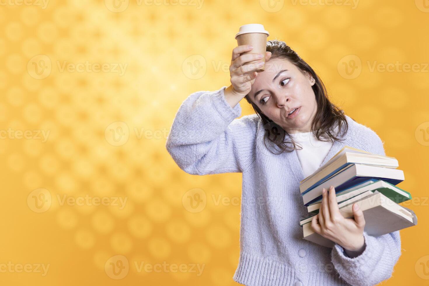 Woman feeling migraine from stress, carrying books for school exam and coffee cup to wake up, studio background. Exhausted student suffering headache from too much homework, drinking caffeine beverage photo