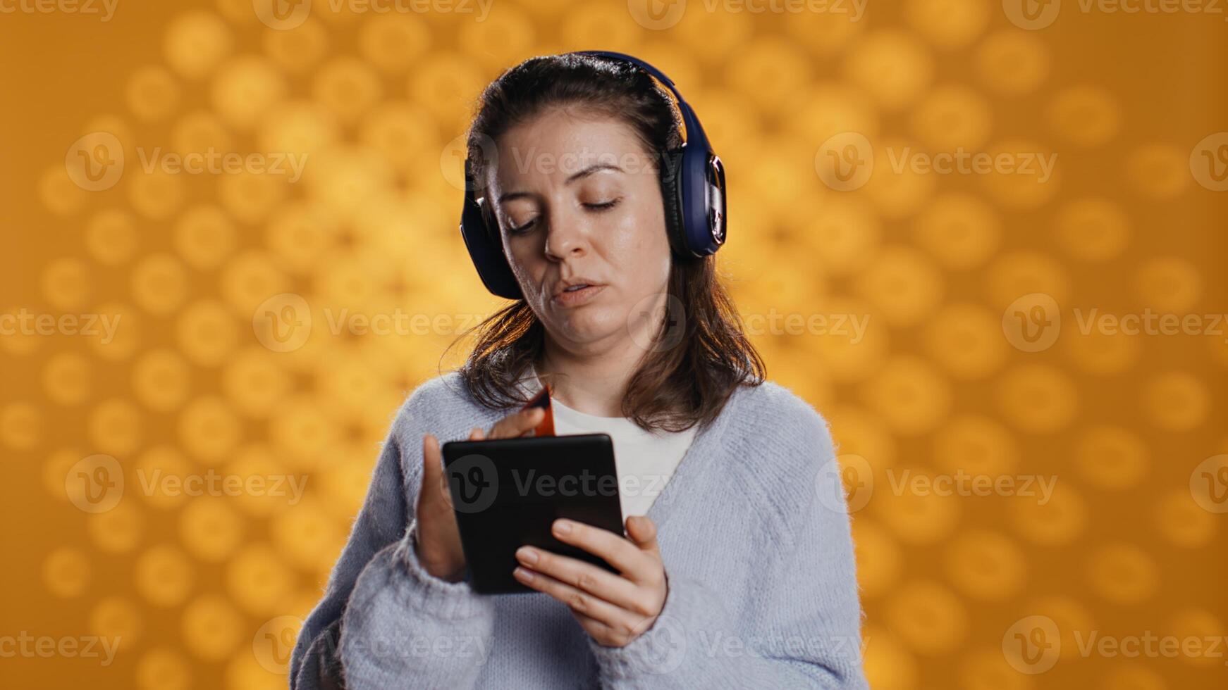 Woman holds ebook reader, listening music and purchasing books online. Person with tablet in hands buying digital novel with credit card, hearing songs in headphones, studio background, camera B photo