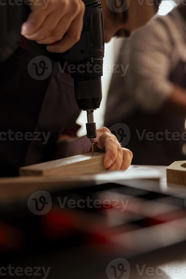Carpenter drilling hole into timber block in studio helped by coworker, close up. Woodworking expert in joinery inserting nail into piece of wood using power drill with apprentice in background photo