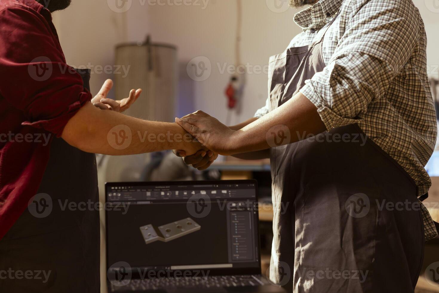 Carpenter meeting with client in studio, shaking hands and talking about commissioned furniture assembling job. Man greeting customer, showing him blueprint on CAD software on laptop photo
