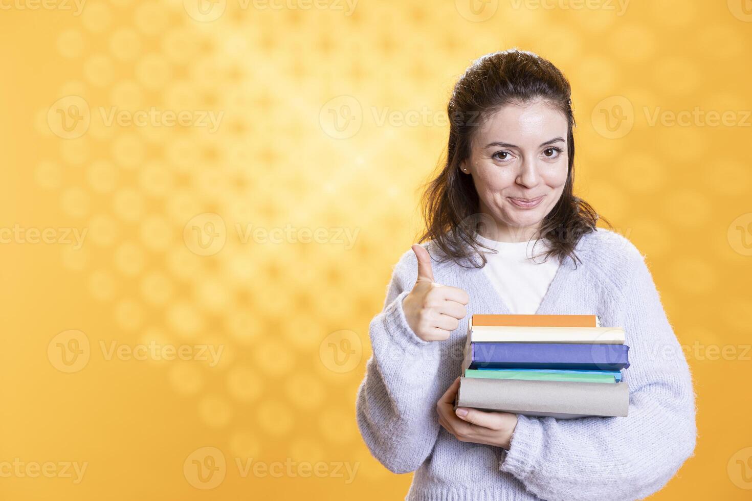 Portrait of happy woman with stack of books in hands showing thumbs up, isolated over studio background. Joyous bookworm holding pile of novels, feeling upbeat, doing positive hand gesturing photo