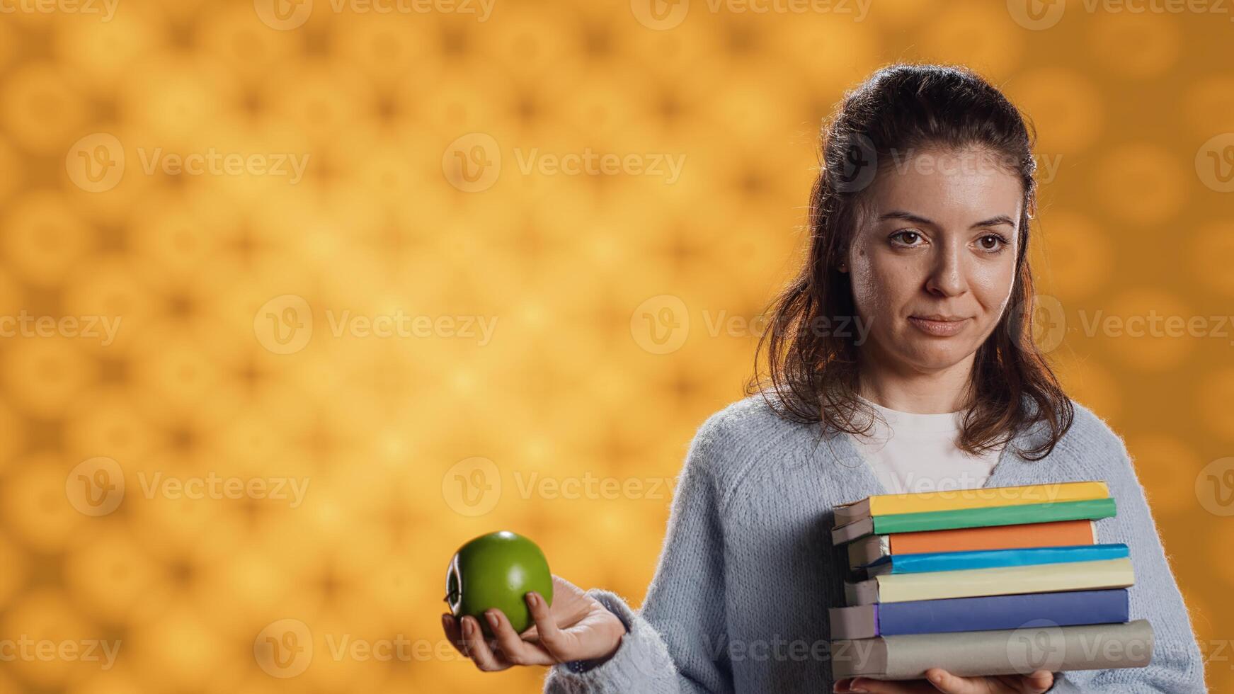 Woman with stack of books in hands throwing apple in air, having fun, living healthy, studio background. Portrait of bookworm holding pile of novels and green fruit, enjoying bio snack photo