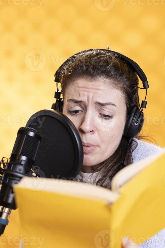 Woman frowns while doing voiceover reading of book to produce audiobook using melodramatic acting. Narrator glowers, using storytelling skills to portray character, recording novel, studio background photo