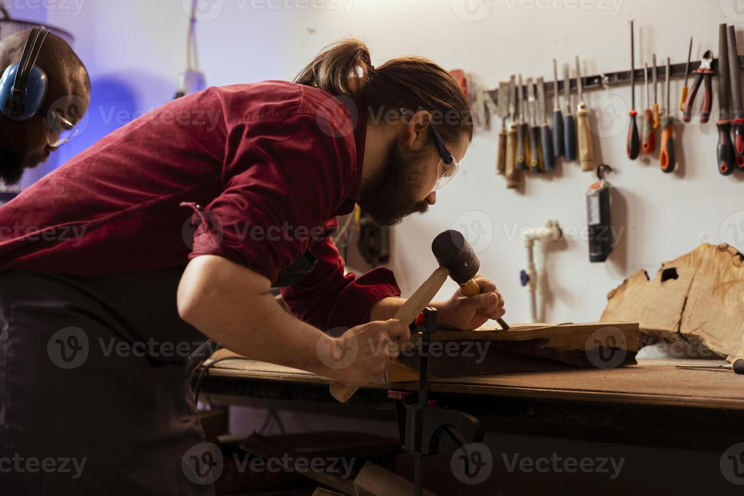 Experienced woodworker shaping raw timber using chisel and hammer in carpentry shop, wearing safety glasses. Woodworking expert shaping wooden objects, making wood sculptures using protective gear photo