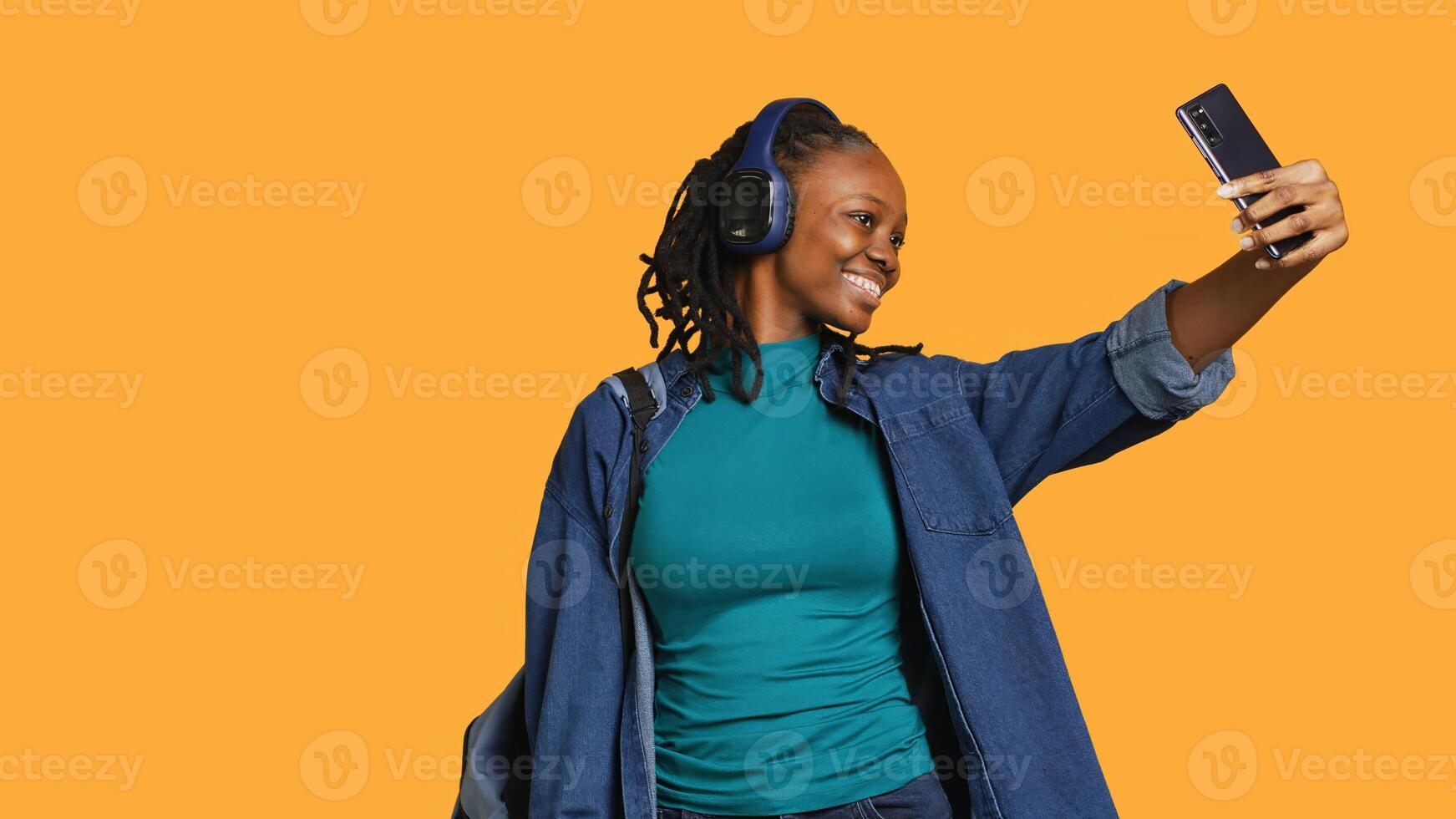 Cheerful woman with headphones using smartphone to take selfies and post them on social media. Happy person taking photos using phone selfie camera, isolated over studio background