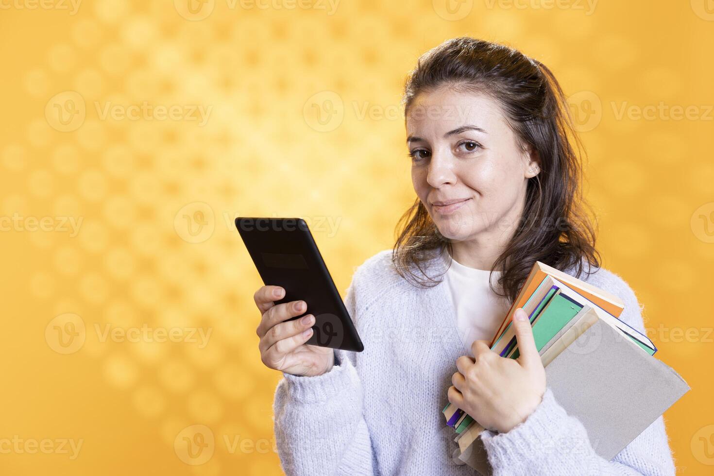 Portrait of smiling woman holding pile of books and ebook reader, conveying joy of reading concept, studio background. Cheerful person with real novels in arms and digital ones on ereader photo