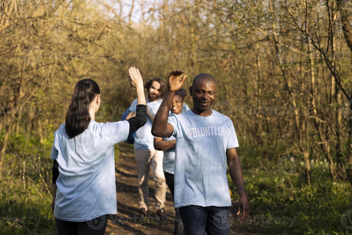 Cheerful satisfied activists sharing high five with each other in the woods, finishing to tidy up the forest ecosystem against pollution. Pleased volunteers showing praise after a job well done. photo