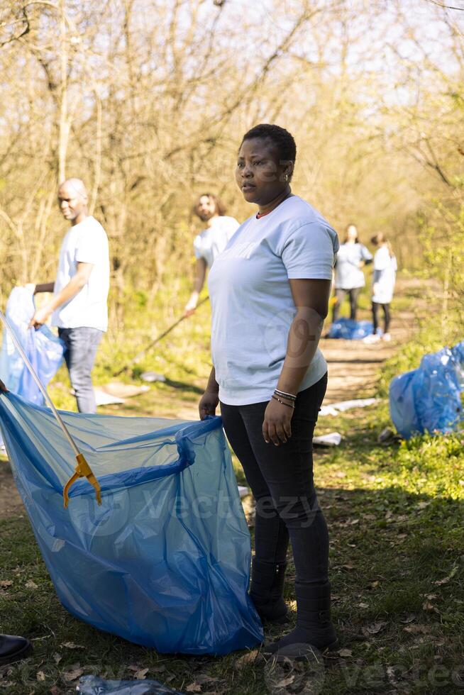 African american woman grabbing trash with a claw tool in the woods, clearing natural habitat of garbage and plastic junk. Activist doing voluntary work against illegal dumping. photo
