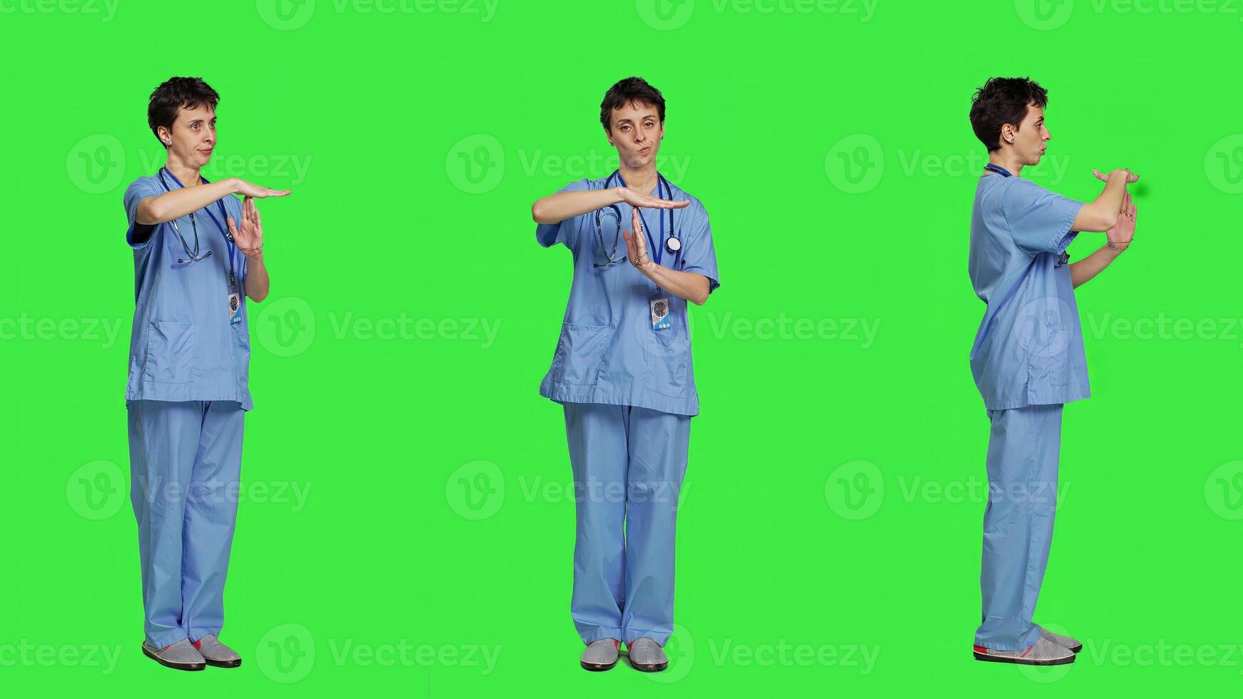 Medical assistant giving timeout symbol against greenscreen backdrop, showing limits and asking for a break. Nurse doing pause or stop gesture while she wears blue scrubs. Camera A. photo