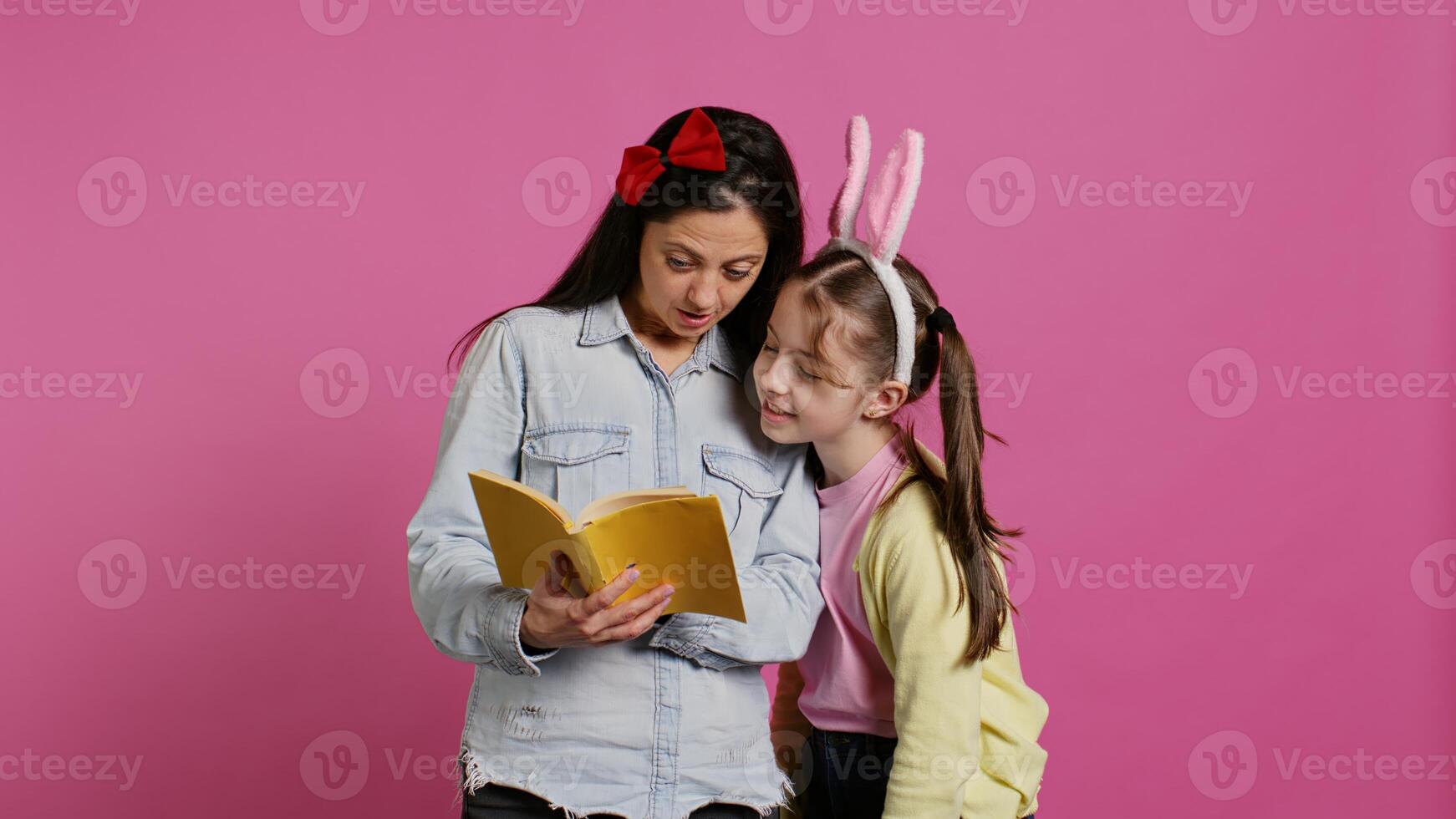 Mother reading fairytale story to her sweet clever daughter, enjoying fiction book with novels against pink background. Cheerful curious schoolgirl with bunny ears listening to her mom. Camera B. photo