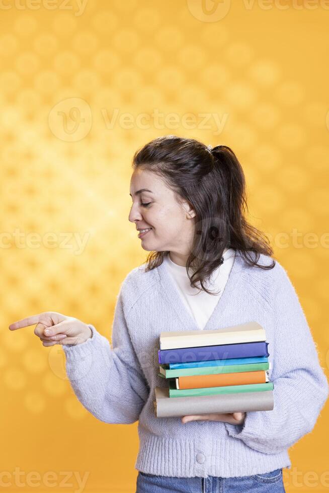 Portrait of upbeat person with stack of books in hands pointing towards empty copy space, isolated over studio background. Cheerful bookworm holding pile of novels, pointing finger off camera photo