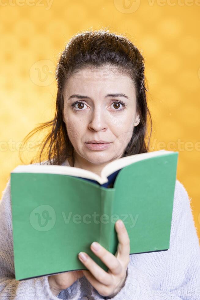 Portrait of woman with worried look on face, appalled by lecture book needed for school exam, studio background. Student reading difficult academic textbook needed to pass classes, feeling anxious photo