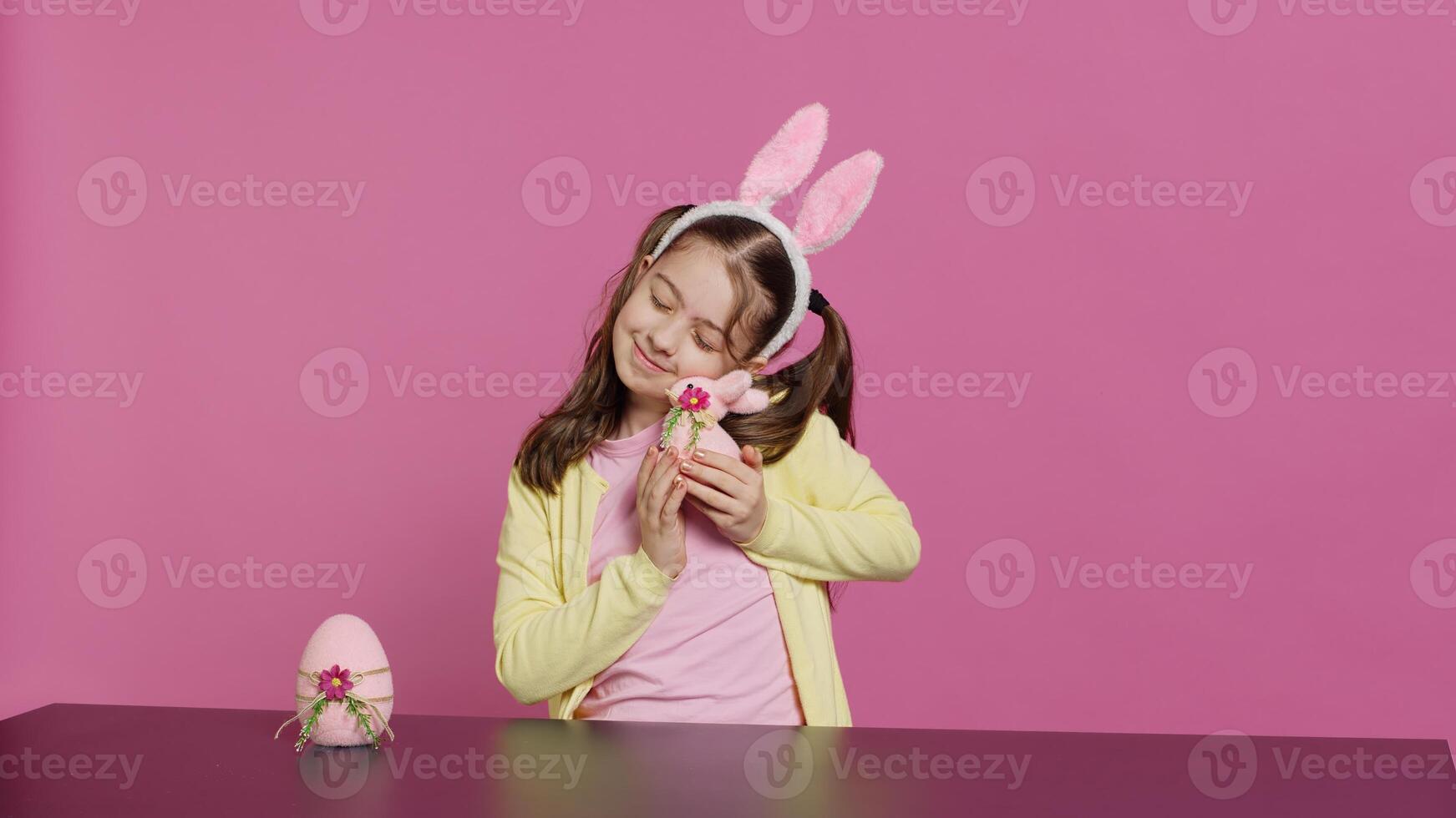 Delighted little girl playing around with lovely easter decorations, creating festive ornaments to celebrate spring holiday festivity. Cheerful kid presenting cute pink rabbit and egg. Camera A. photo