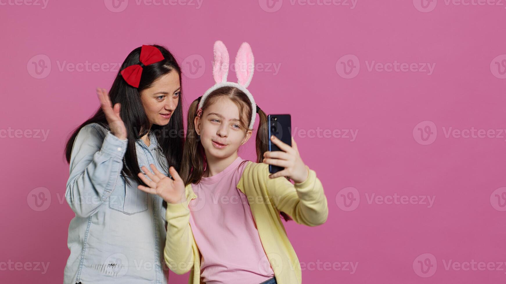 Little schoolgirl and her mom talking on videocall meeting in studio, waving at webcam and chatting with their family. Cute preteen and mother using smartphone for a teleconference chat. Camera A. photo