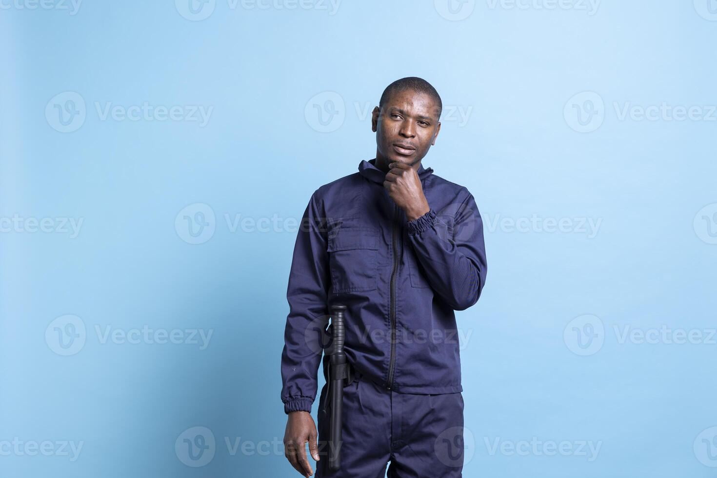 Thoughtful security personnel with uniform and baton thinking of ways to better protect the premises, blue background. Pensive guardian working on safeguarding and securing, patrol service. photo