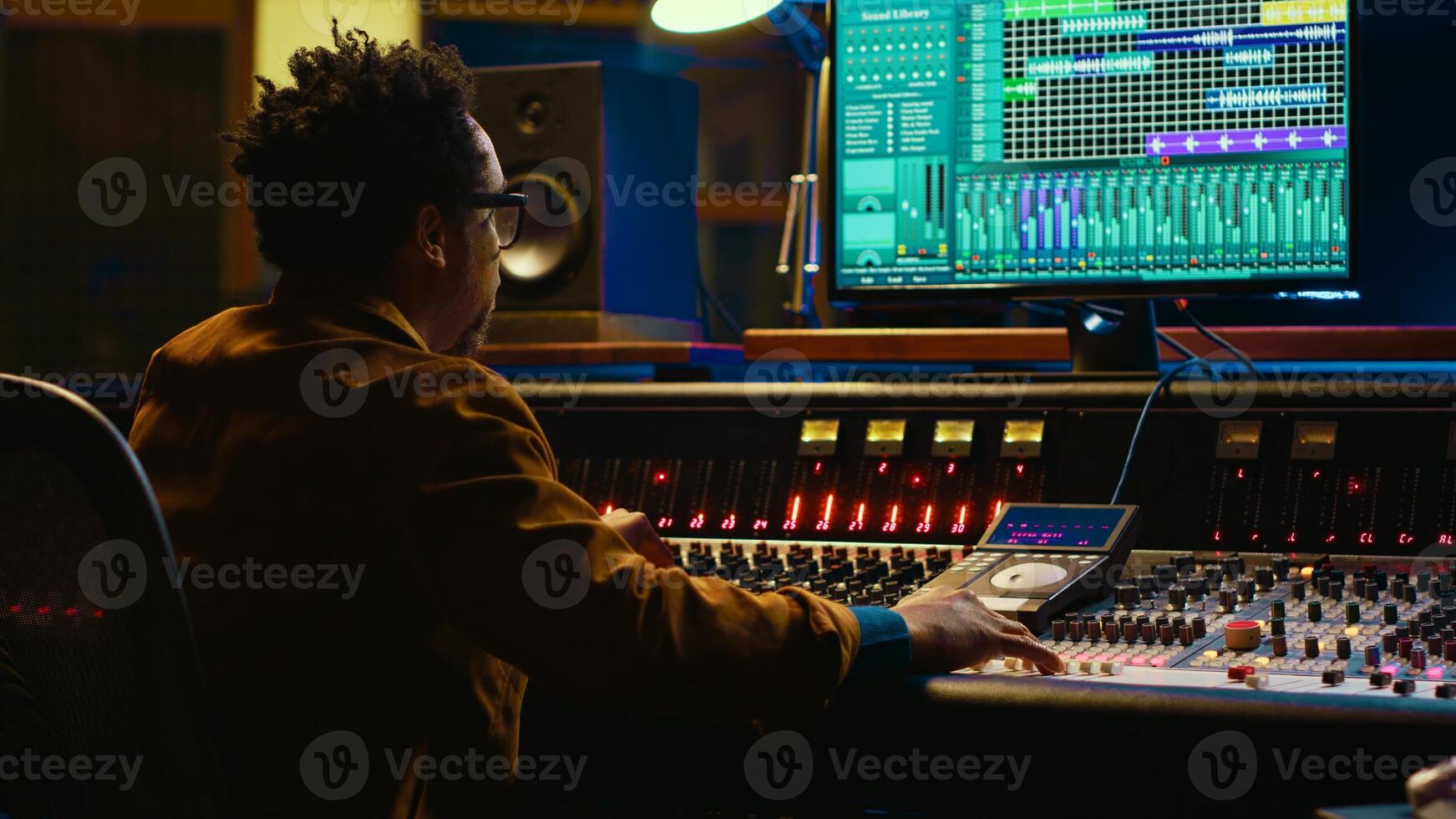 African american sound expert operates on audio console with moving faders and colored meters, editing in professional studio control room. Producer works on album, recording hit song. Camera A. photo