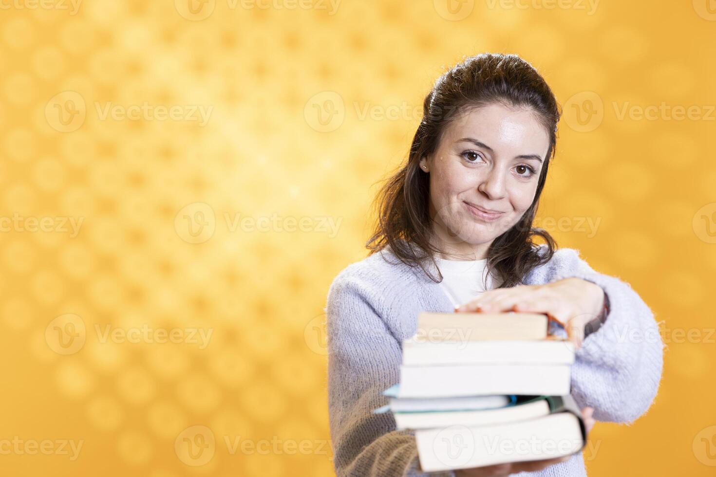Woman holding stack of books, pointing it towards camera, isolated over yellow studio background. Portrait of smiling bookworm lady presenting stack of novels, urging people to read photo