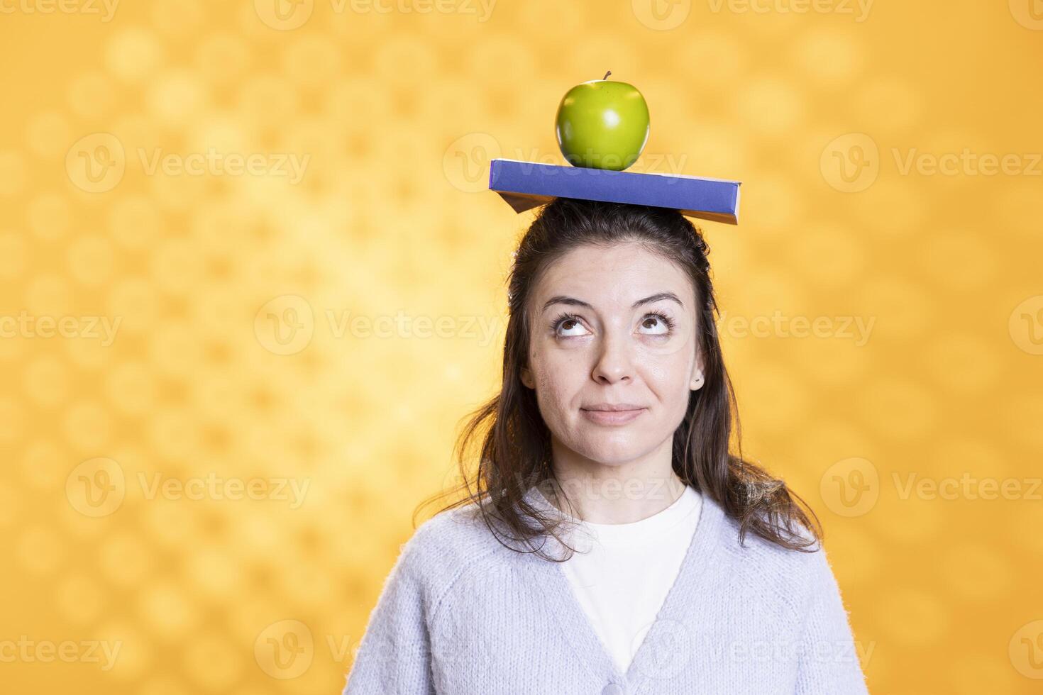 Woman with book on head and fresh apple acting zany, portraying living healthy, isolated over studio background. Bookworm wearing novel and green fruit as hat, enjoying life, goofing around photo