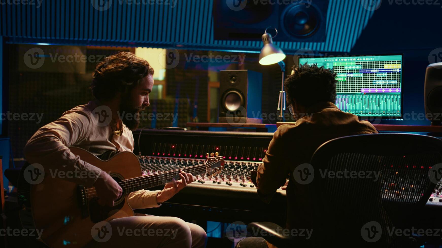 Composer musician recording a new song on his electro acoustic guitar in professional studio, using mixing console and amplifier in control room. Young artist composing new tracks. Camera B. photo
