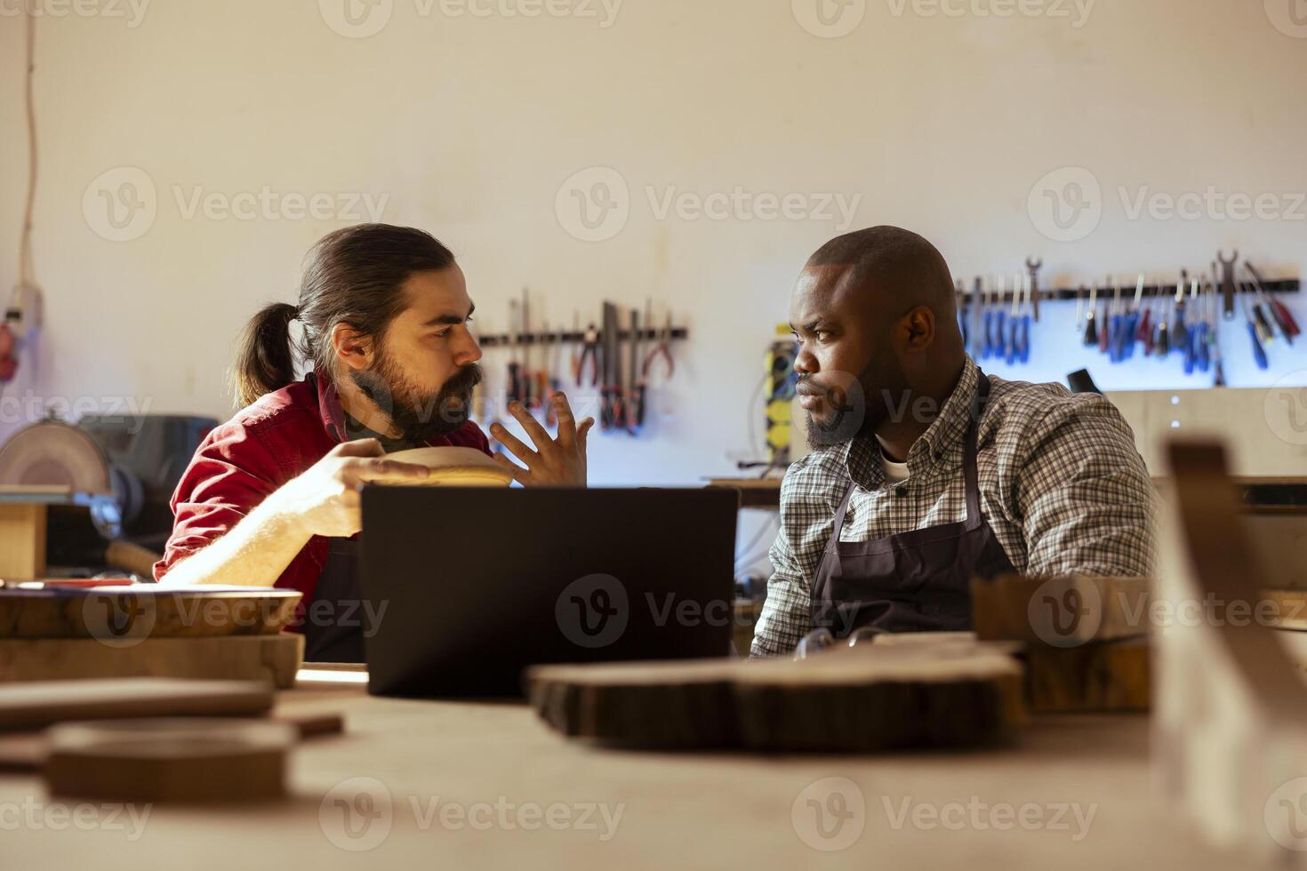 Woodworker looking over blueprints on laptop, brainstorming with african american apprentice. Manufacturer and coworker discussing furniture assembly schematics on notebook in studio photo