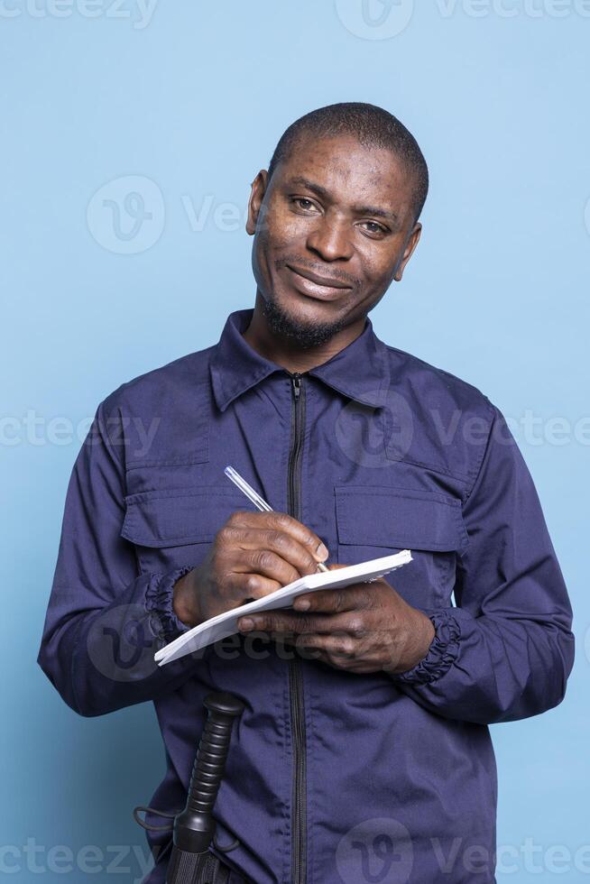 Security officer in work uniform writes important details on notebook, posing against blue background. Smiling proud mall guard taking notes of information to keep evidence of responsibilities. photo