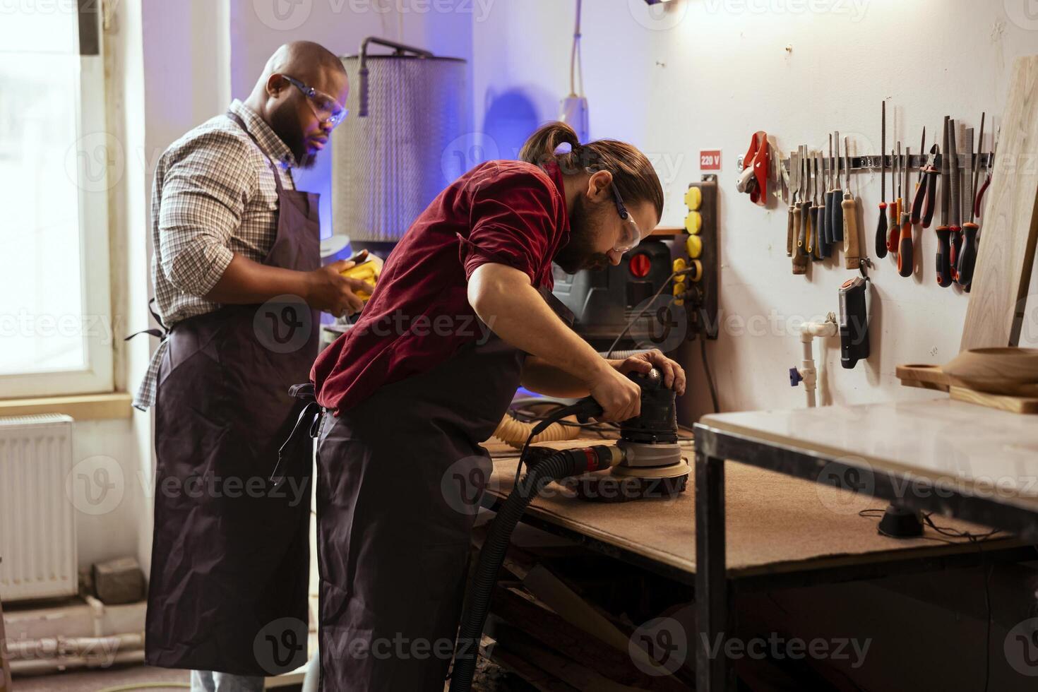 Master carpenter in joinery watching apprentice using orbital sander on lumber. Team in woodworking shop using angle grinder to create wooden decorations, refining wood objects photo