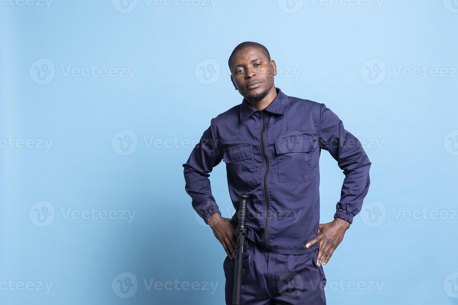 Security guard showing his authority by posing with hand on his hips against blue background, confident armed bodyguard. Male agent feeling proud in his work uniform while on duty. photo
