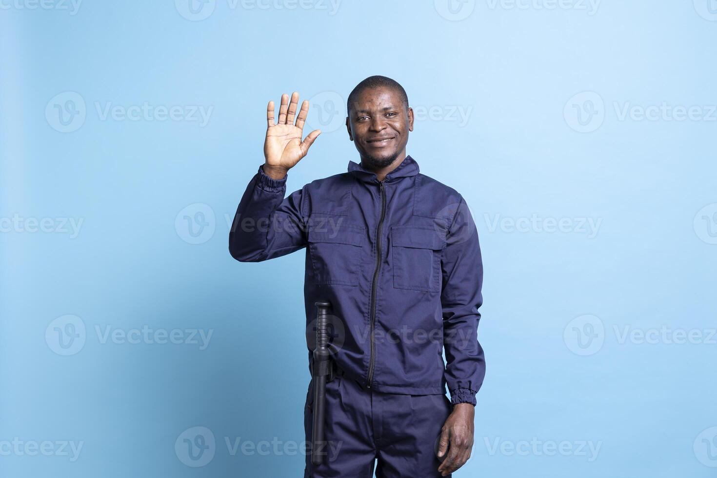 Cheerful african american security guard waving at the camera, greeting people and saying hello. Young defense protection agent safeguarding property, serious responsibility against blue background. photo