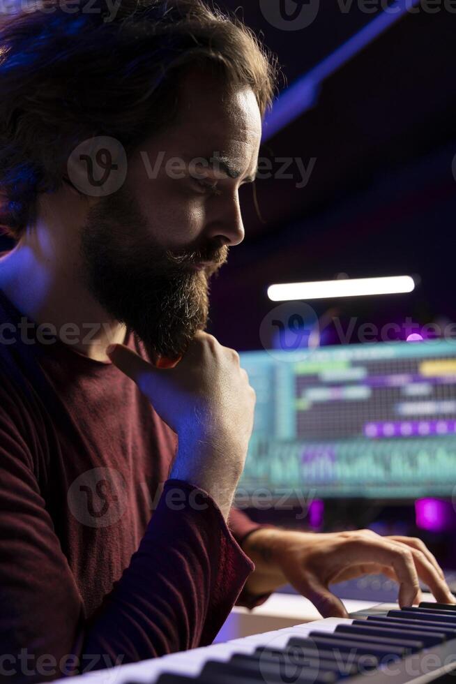 Artist thinking of new piano notes combinations to play on his electronic keyboard, recording music with daw software in home studio. Musician searching for inspiration in playing synthesizer. photo
