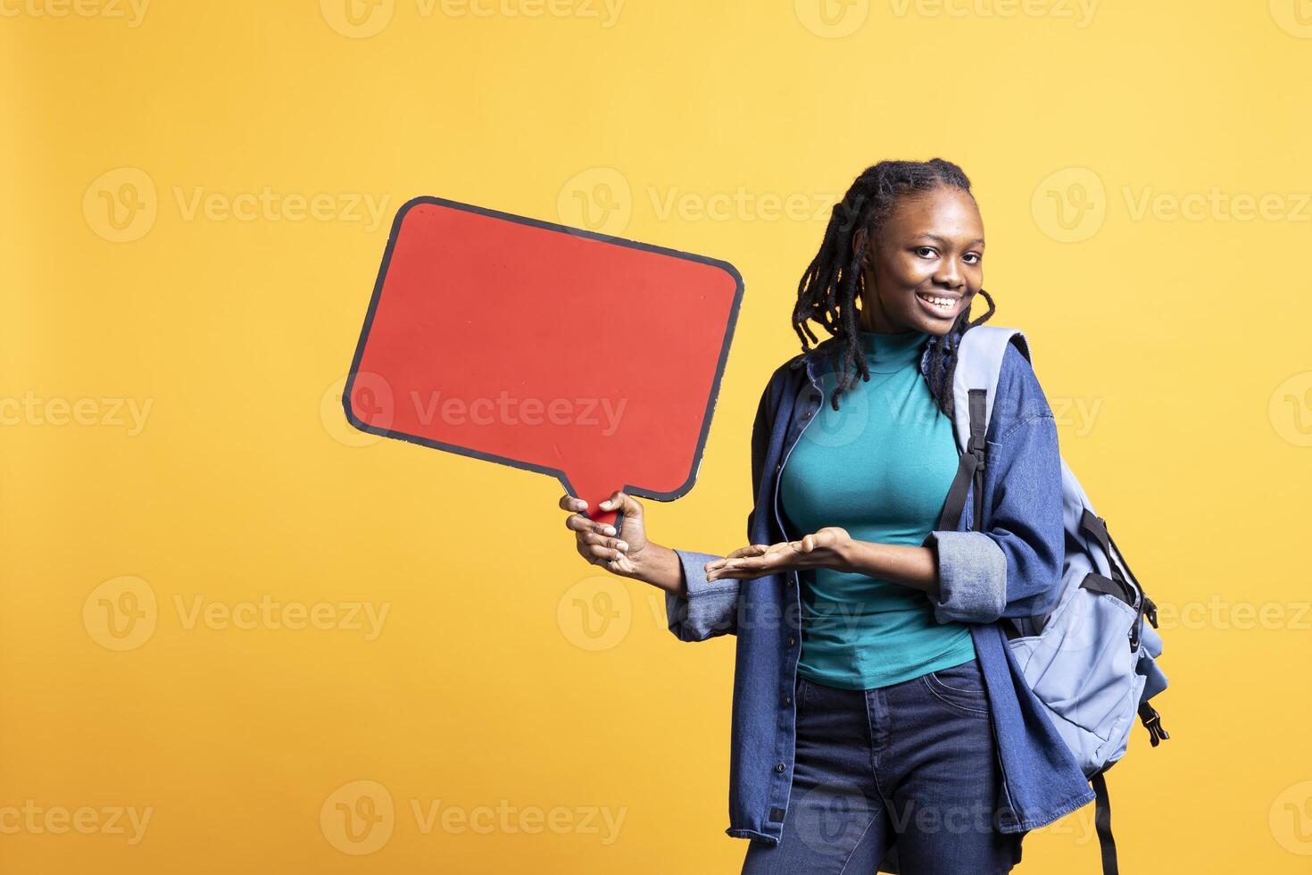 Radiant BIPOC student with backpack holding red speech bubble sign of empty copy space. Upbeat woman presenting thought bubble cardboard, advertising concept, studio backdrop photo