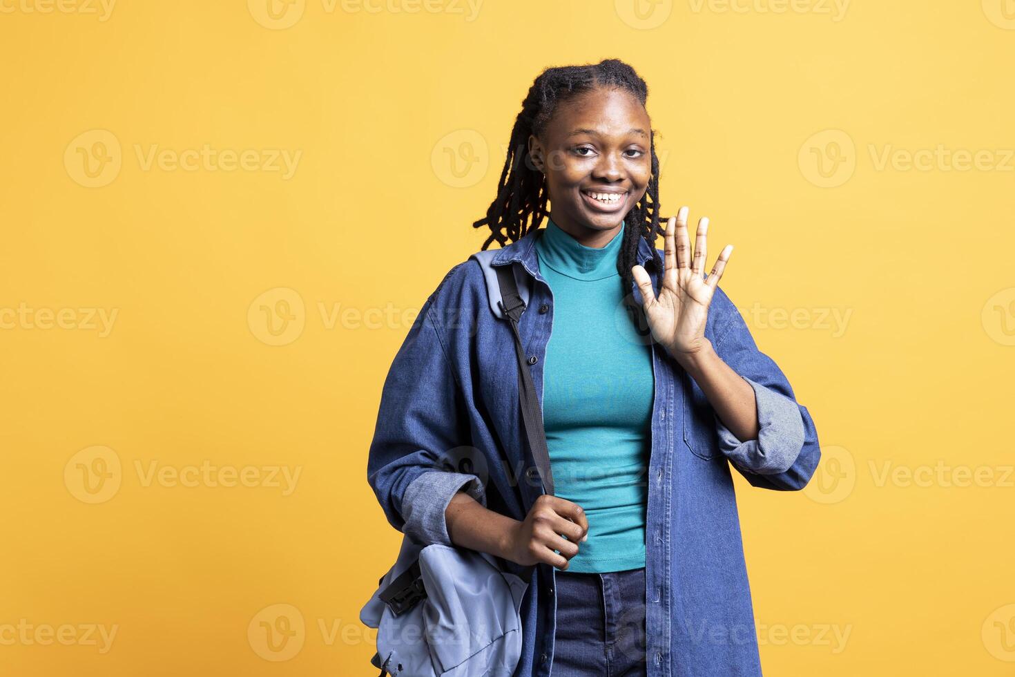 Portrait of happy woman holding backpack, waving hand, gesturing, isolated over yellow studio background. Cheerful student with school rucksack doing greeting hand gesture photo