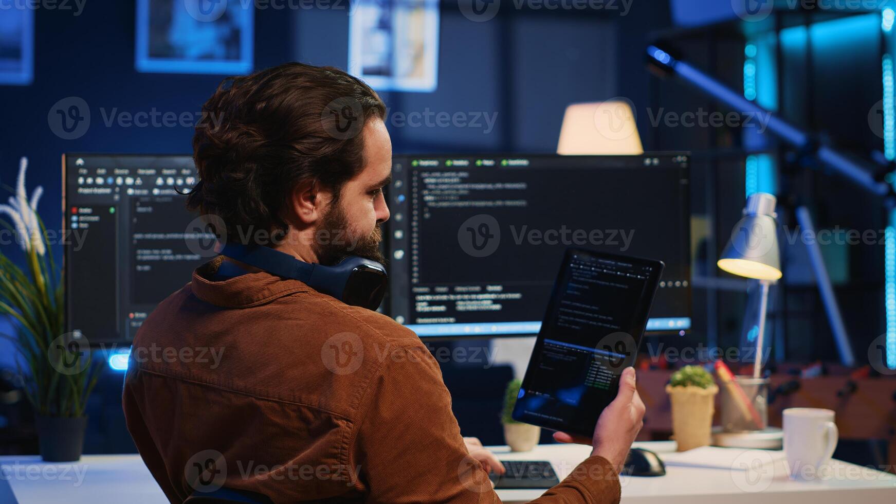 Software developer programming in home office, crosschecking lines of code between tablet and PC screen. IT admin holding portable device, using it as reference while coding on computer, camera A photo