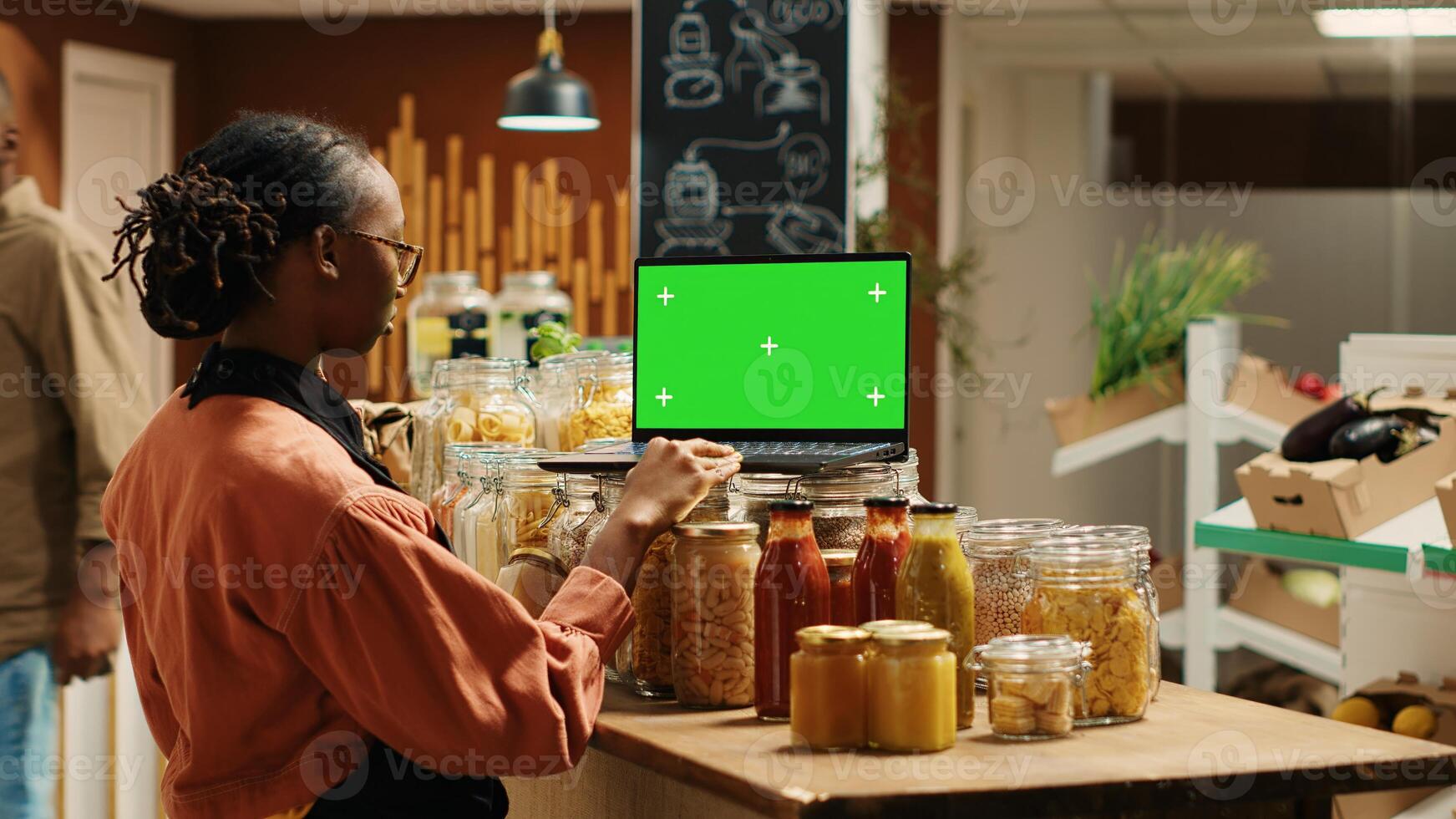 Woman merchant uses greenscreen on laptop and arranges products on stall, sorting bulk items in recyclable jars. Farmers market worker navigating on pc showing isolated chromakey template. Camera 1. photo