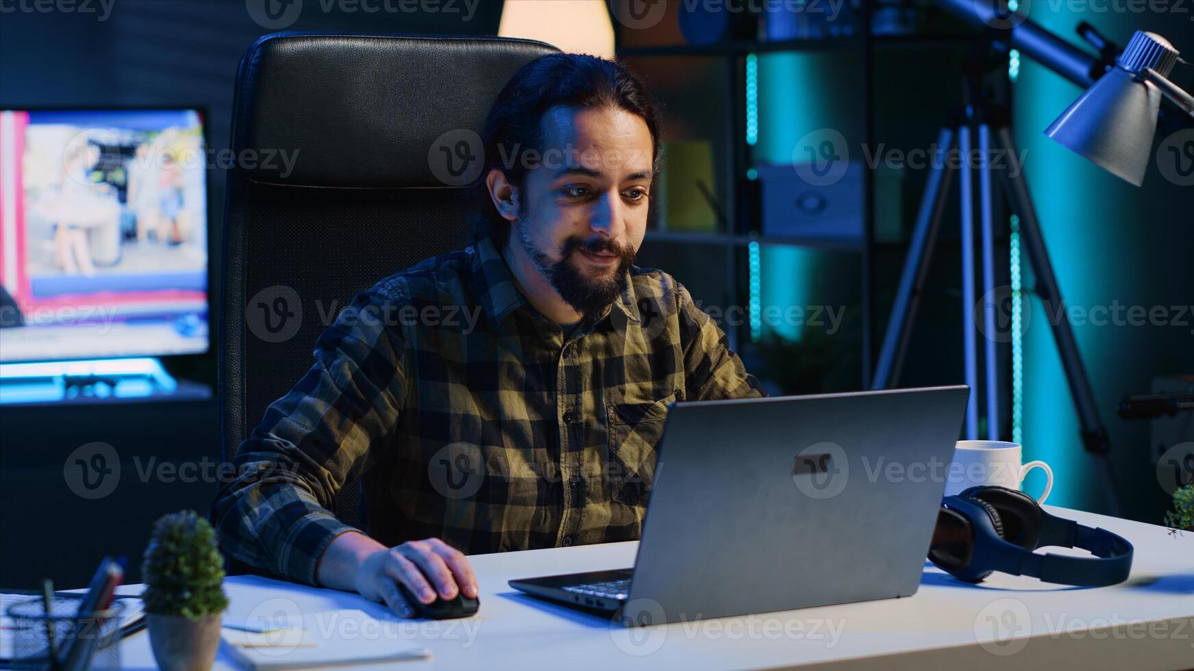 Freelancer in neon lit home office sitting at desk and typing on laptop keyboard. Smiling remote employee using electronic device to do customers relations job online, answering inquiries, camera A photo