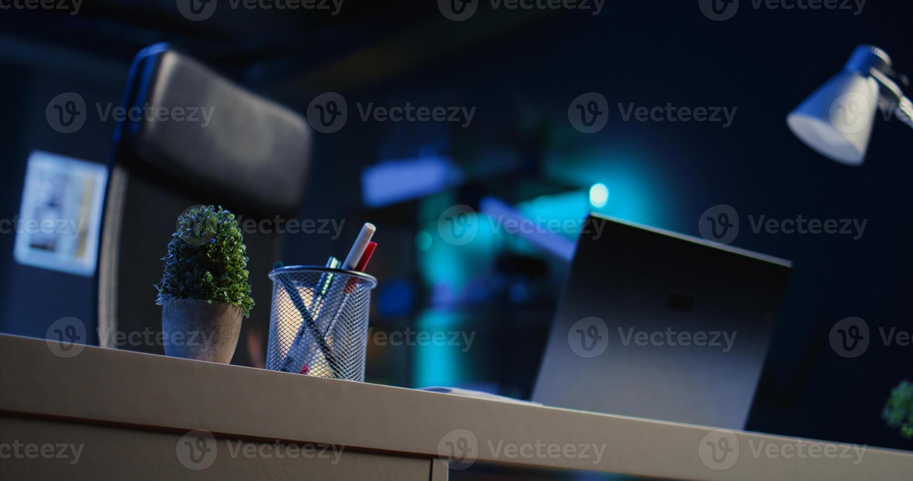 Office desk filled with productivity items in empty home interior illuminated with neon lights at night. Laptop on table and other work related objects in apartment room, camera B close up shot photo