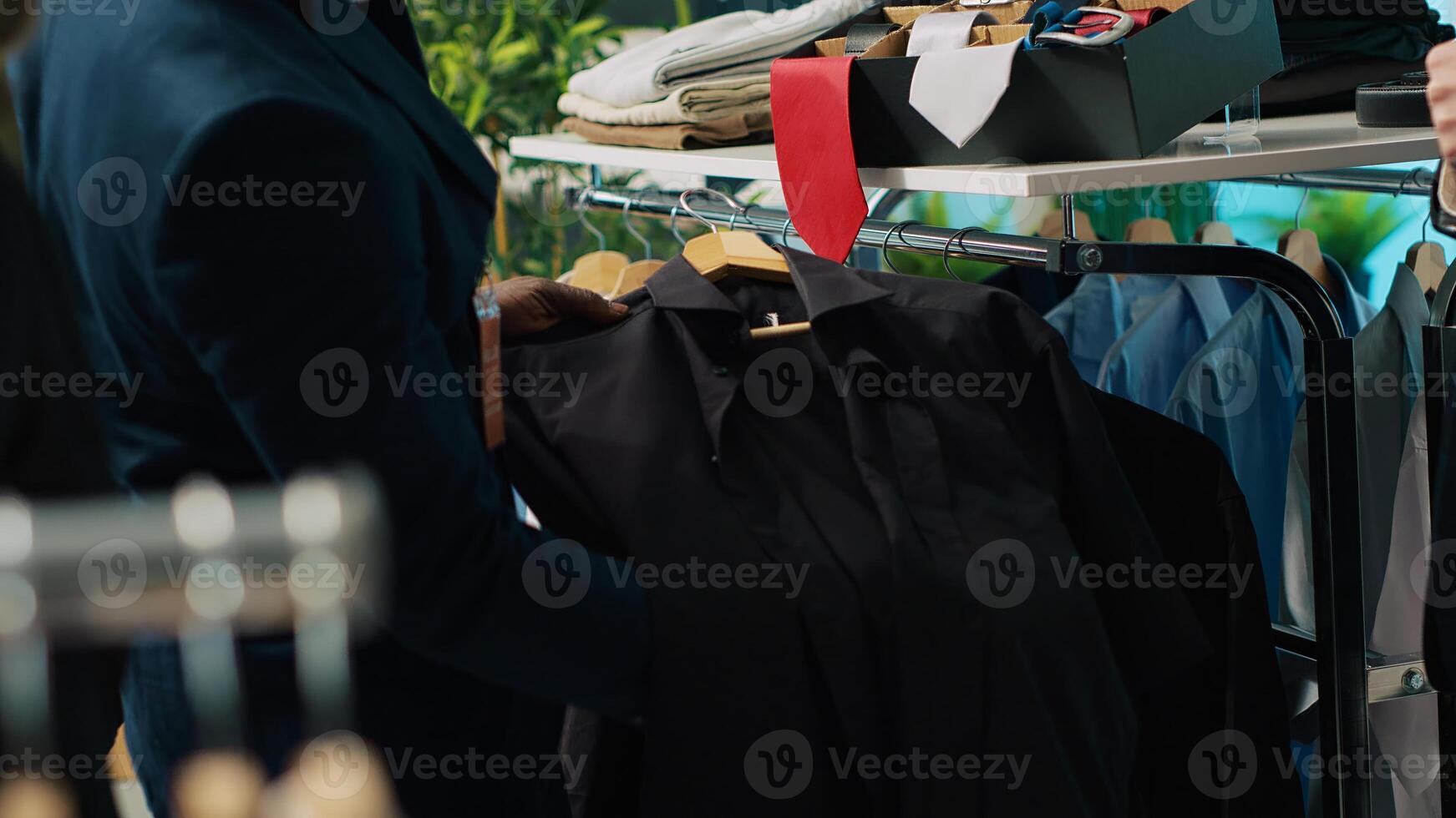 African american worker showing a black shirt and red tie to client, assisting him to buy the perfect outfit for an elegant occasion. Professional stylist gives advice to buyer at store. Camera A. photo