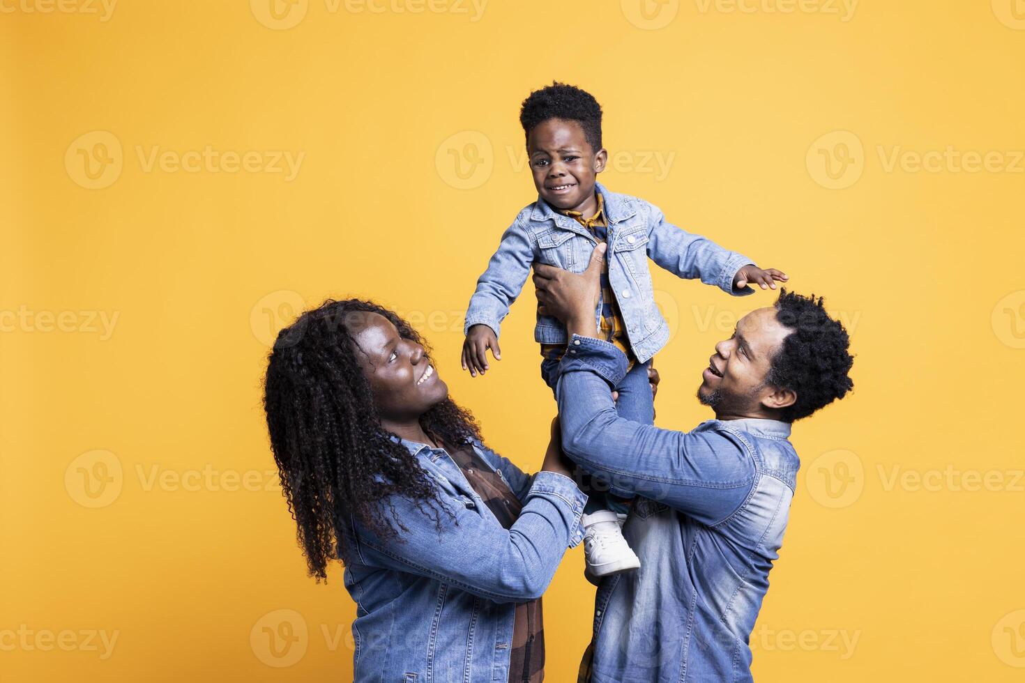 Loving african american parents lifting their cute small son in studio, posing in front of the camera. Proud happy couple bonding with their adorable toddler against yellow background. photo