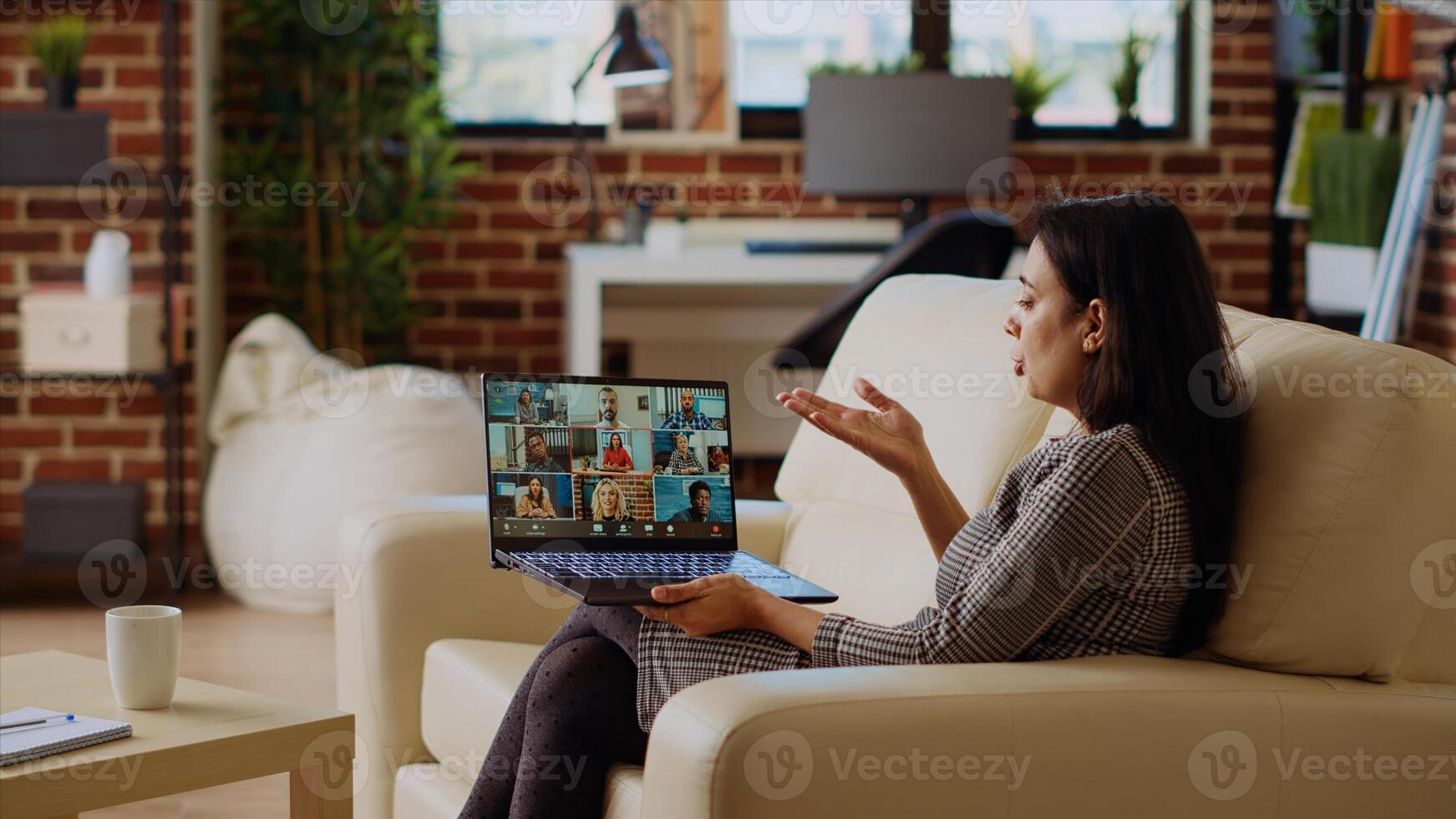 Teleworking team leader sitting on couch in cozy apartment, chatting with coworkers during teleconference meeting. Indian business manager at home holding online videocall with workers, camera B photo