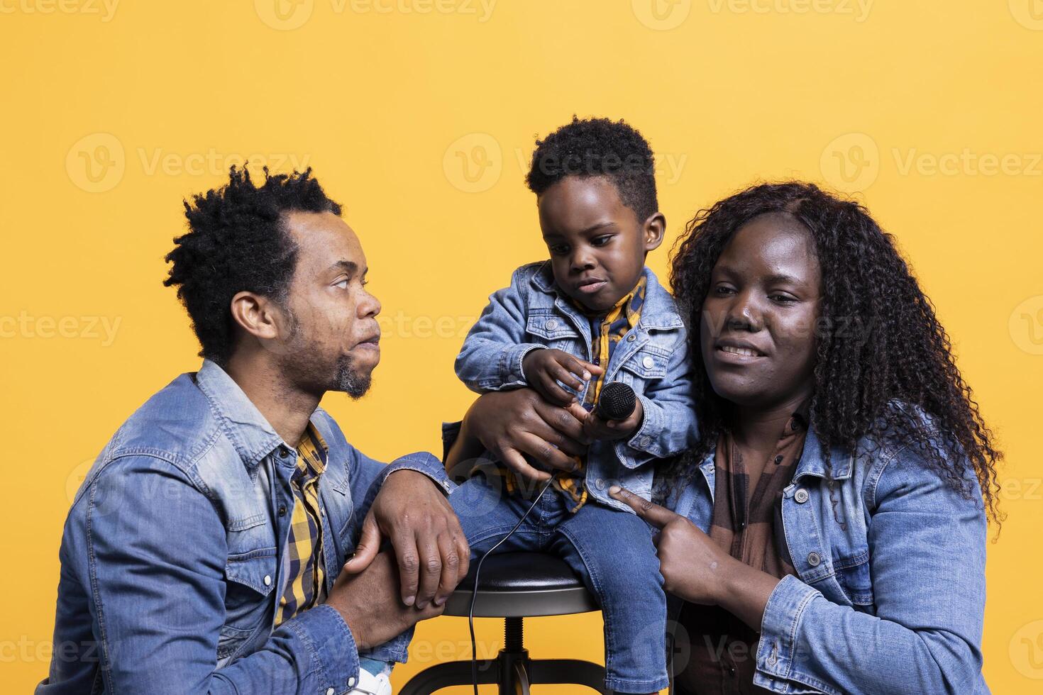 Adorable small kid playing with a microphone in front of the camera, having fun with his young parents against yellow background. Happy proud african american mom and dad sing with son. photo