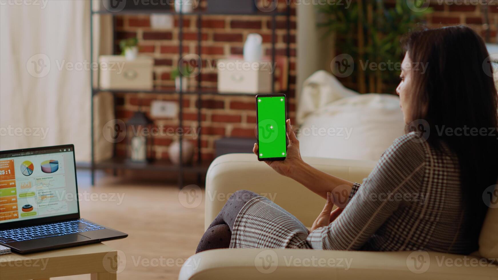 Remote worker looking at isolated screen cellphone while optimizing market performance charts. Indian woman checking online business analytics statistical data metrics on chroma key phone and laptop photo