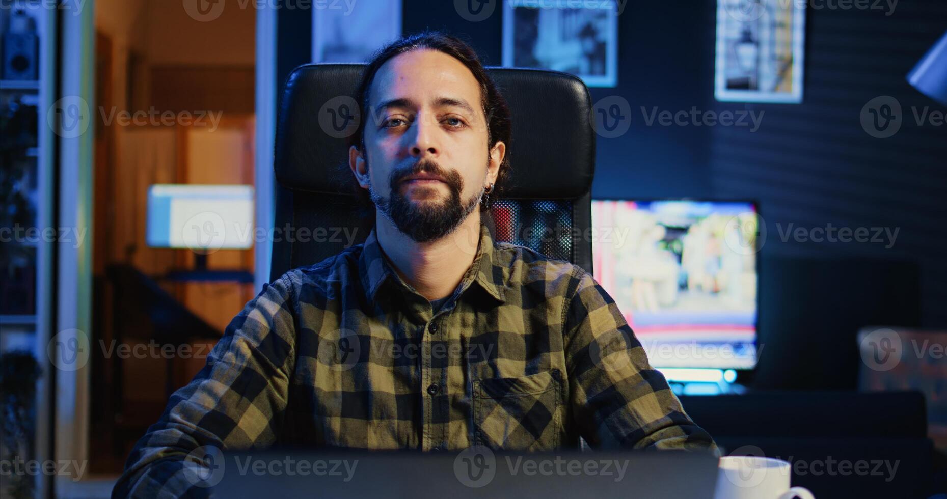 Smiling caucasian man sitting on chair at home office desk in neon illuminated apartment late at night. Cheerful person in RGB lit living room enjoying leisure time, camera B photo