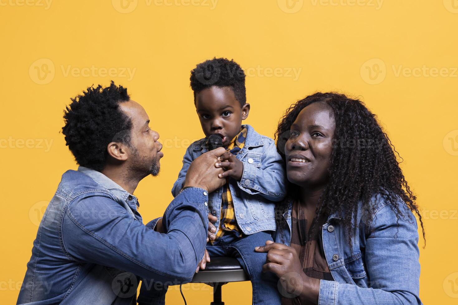 Cheerful adorable small child singing a song on microphone in studio, parents smiling and dancing on his music. Proud mother and father having fun with their happy son on camera. photo