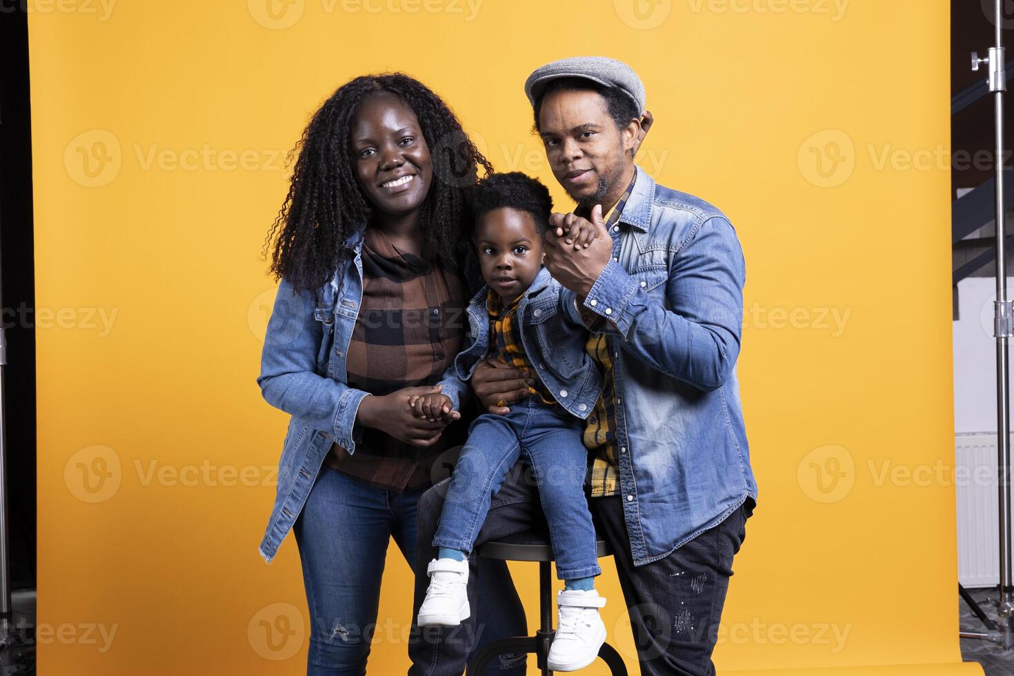 African american joyful family posing together against yellow background, happy couple hugging their little toddler. Smiling young mother and father showing affection to their kid on camera. photo
