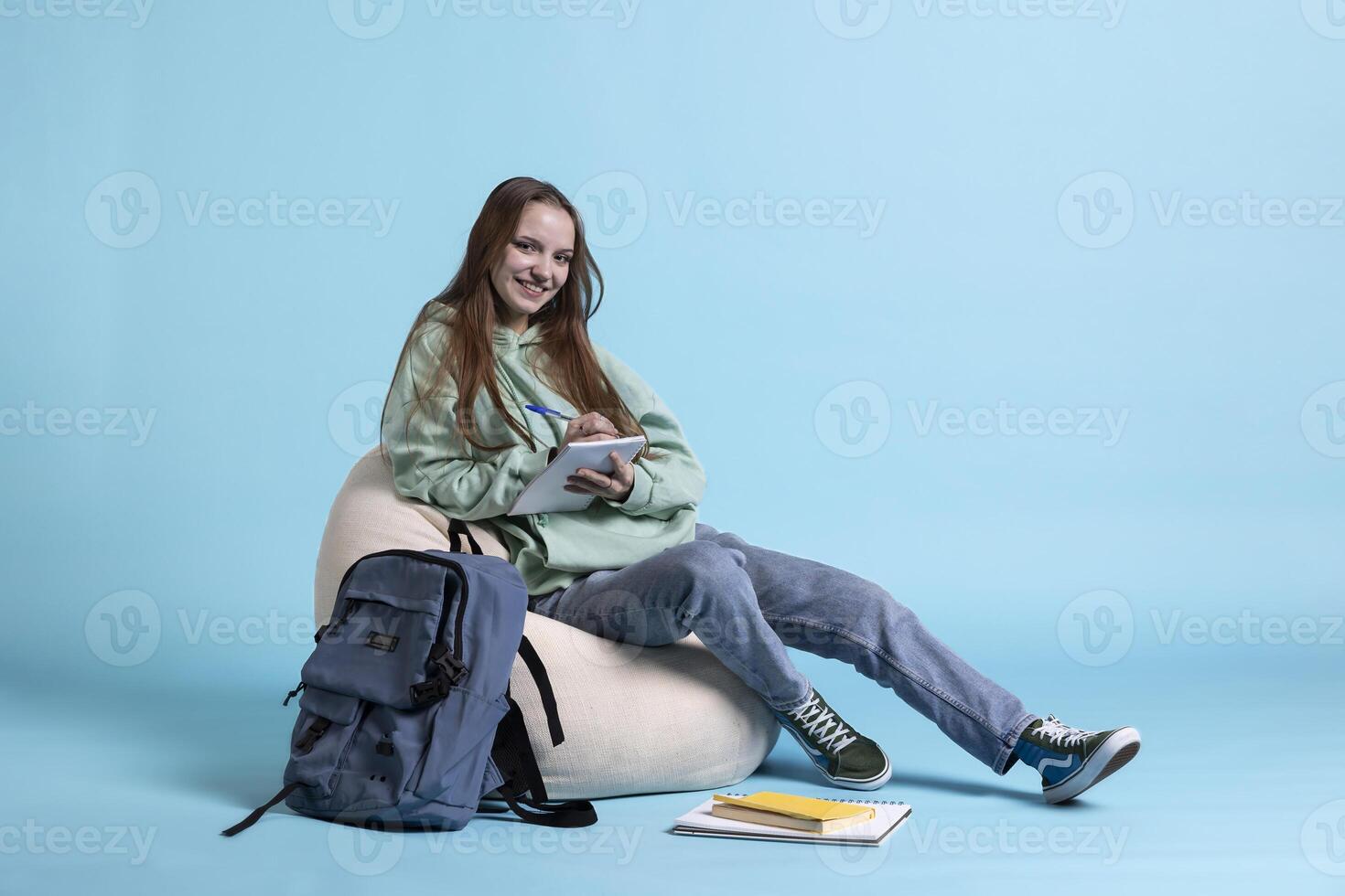Portrait of radiant teenager sitting on beanbag and writing on notebook, studio backdrop. Upbeat schoolgirl with notepad in arms, preparing homework for high school classes photo