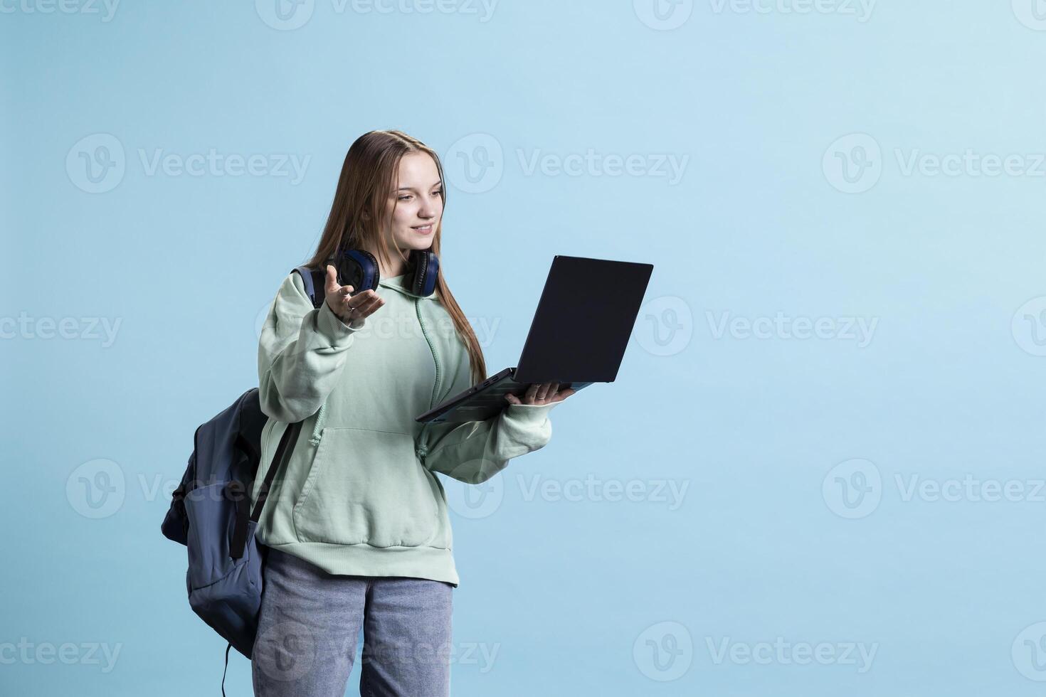Upbeat woman scrolling on laptop to check social media feed, standing up. Carefree teenager browsing online websites on notebook device, isolated over blue studio background photo