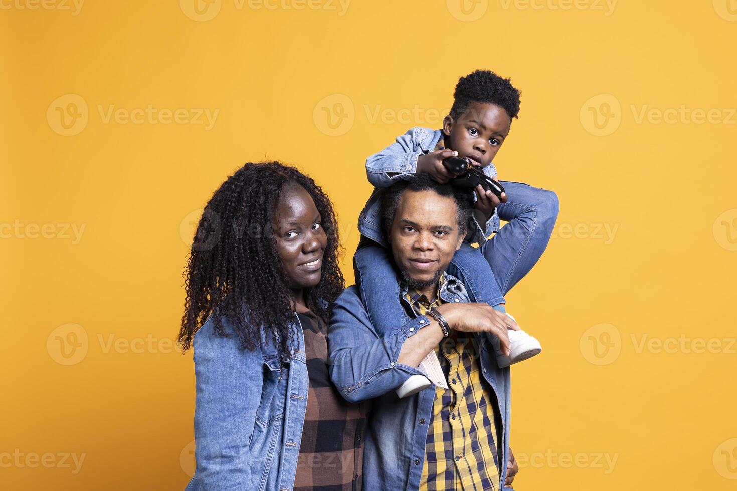 African american dad carrying his toddler on his shoulders against background, sweet kid having fun on camera with a joystick in his hand. Joyful young mother and father bonding with their son. photo