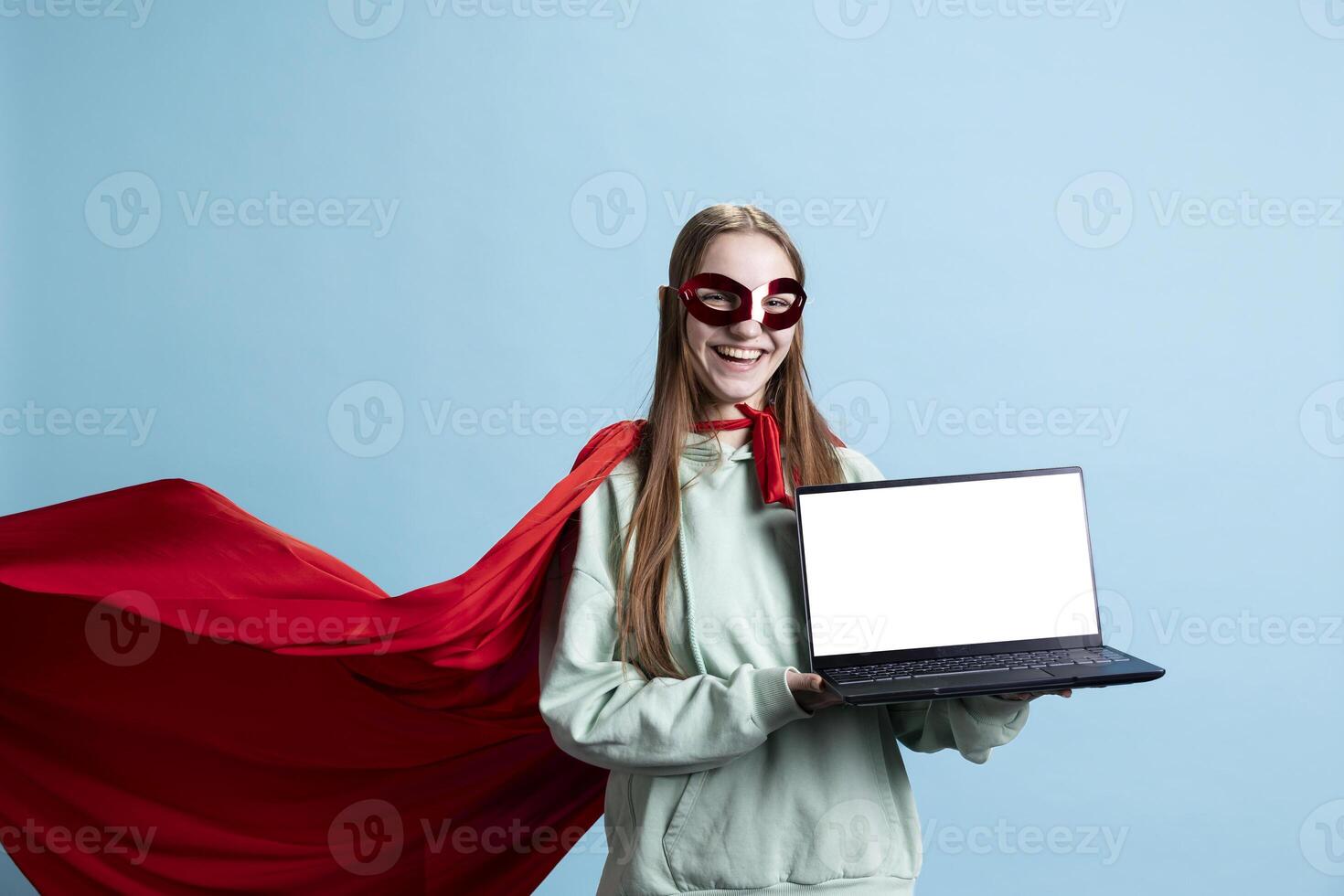 Portrait of happy teenager with mask laughing while holding mockup laptop, portraying superhero, studio backdrop. Jolly heroic person posing as hero character in costume with isolated screen notebook photo
